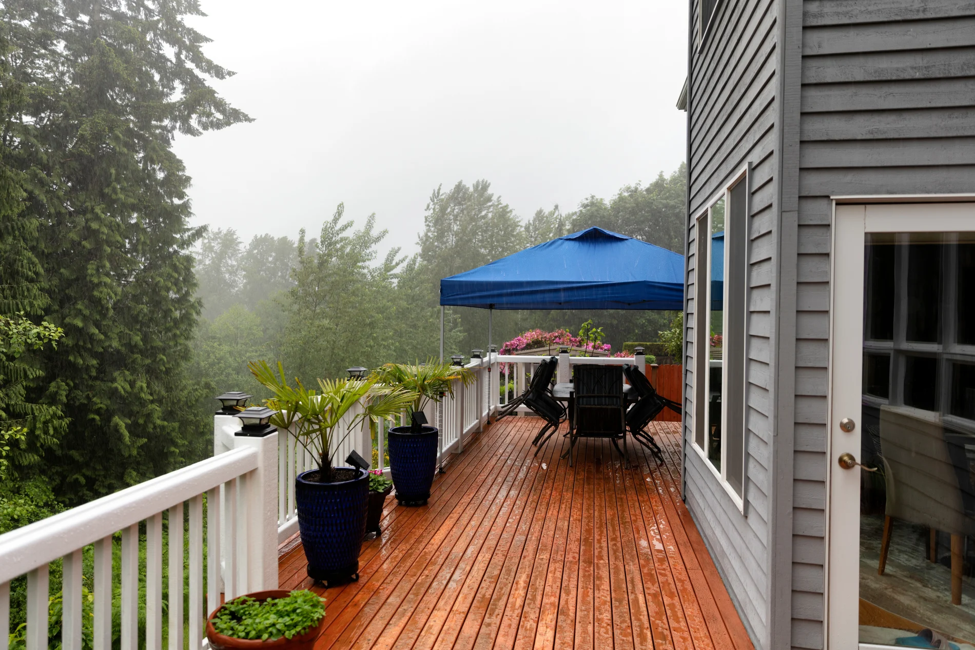 The side of a two-story home, showing a section of a large wood deck with white railing, with rain coming down in buckets and the tops of the trees in the background bending to the wind.