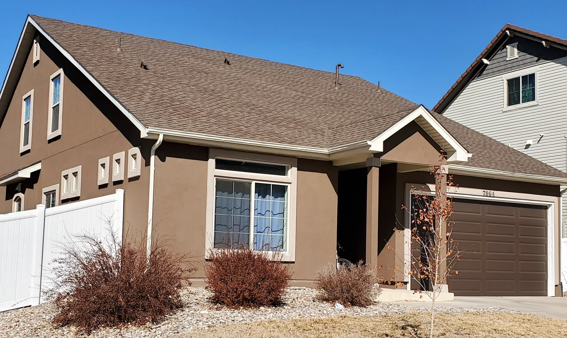 A single-story brown stucco house with white windows, off-white frames, a brown garage door, and a brown shingle roof under a bright blue sky.