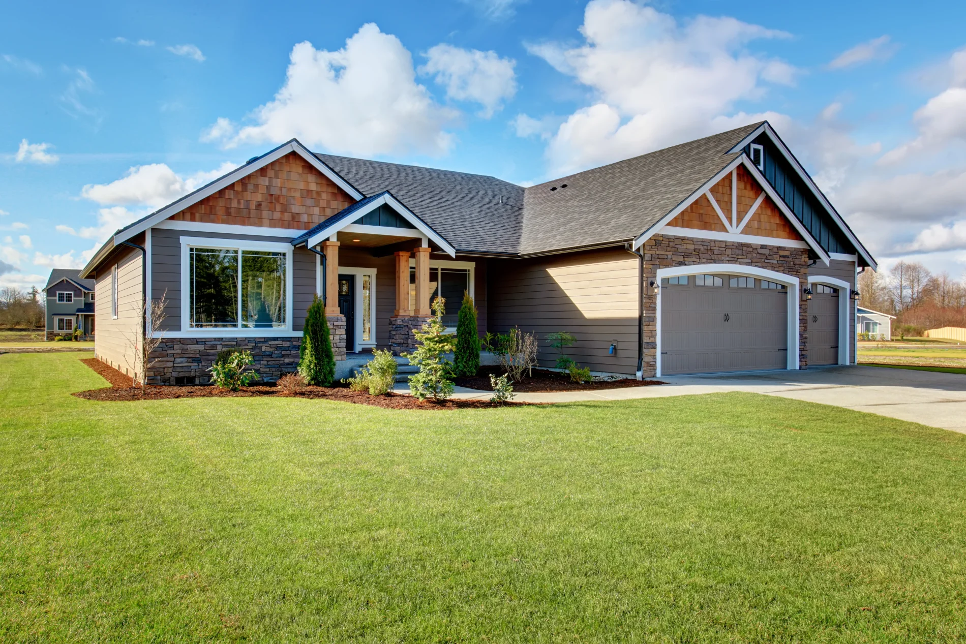 A  green manicured lawn with a beautiful single-story, single-family home with an asphalt shingle roofing system, white fascia, dark soffit, different kinds and colors of panel siding, two garage doors, stone veneers around the front of first garage and at the base around the home’s entrance, white windows and window frames, and simple landscaping.
