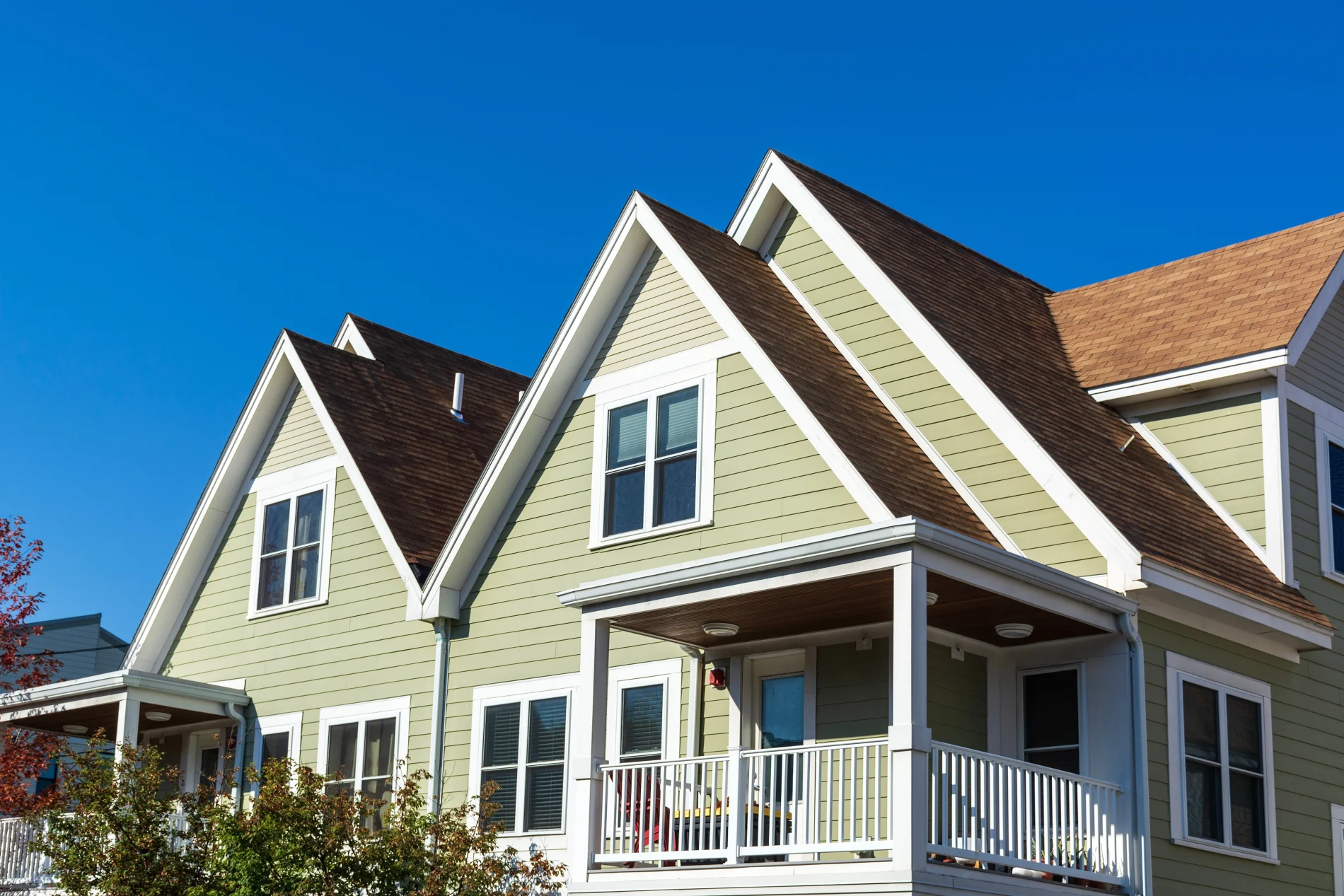 A large elegant family home with light green horizontal panel siding, multiple gable roofs, white fascia and soffit, white windows and window framing, as well as a small covered porch with off-white tiers and gutter system.
