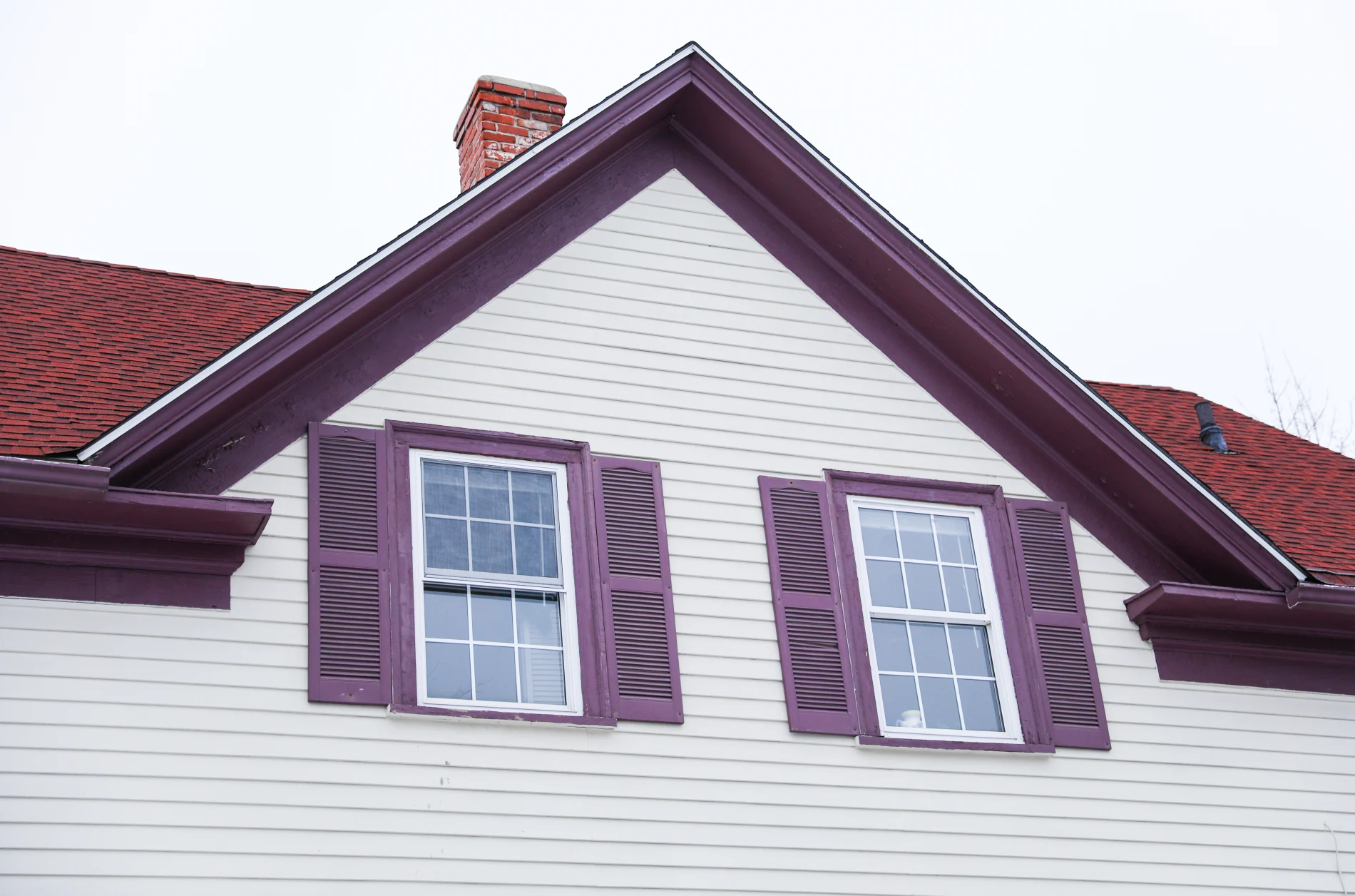A close-up view of the upper floor of a white house with two white windows in teal frames, teal shutters, and teal soffit under a brown shingle roof and the top of a brick chimney.