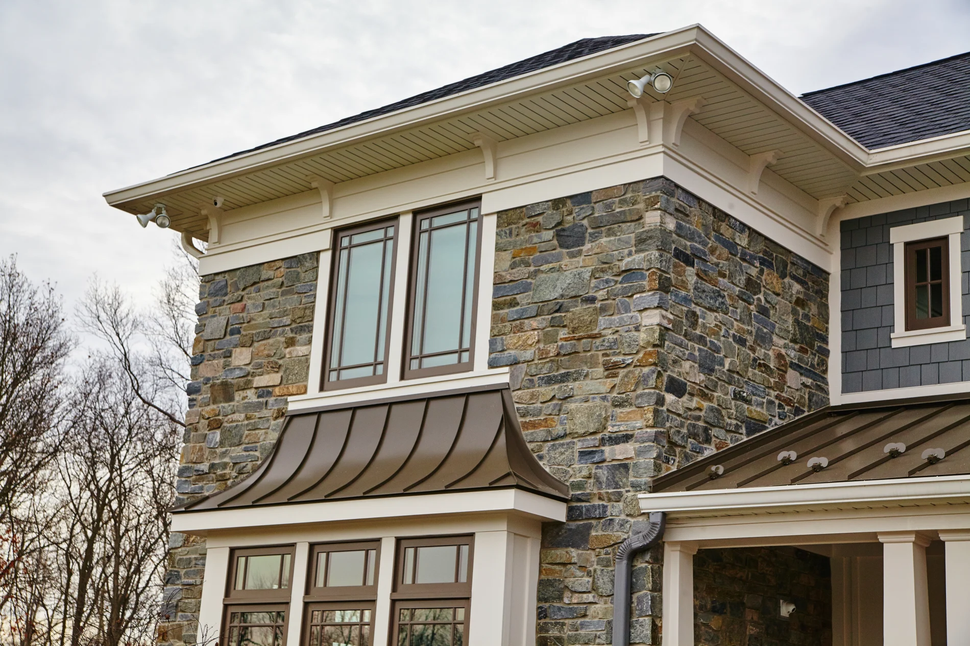 A close-up view of the left side of a two-story house with stone, blue siding on the rest, red metal roofing over the first floor, and a shingle roof on top.
